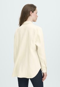 Woman with long brown hair wearing a loose, cream-colored long-sleeve shirt and dark jeans, facing sideways against a plain light background.