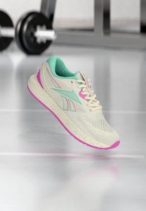 Hvid løbesko med turkis og pink accenter svævende over et fitnessgulv, fitnessvægte sløret i baggrunden.