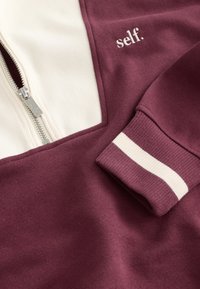 Sarkana rāvēja džemperis ar krēmkrāsas paneli, ar ribotiem aprocēm un izšūtu "self." logotipu. Gluda tekstūra un sportisks dizains.