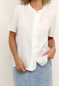 Blouse blanche à manches courtes avec un tissu texturé, un devant froncé et une fermeture par boutons. Portée avec une jupe en denim bleu.