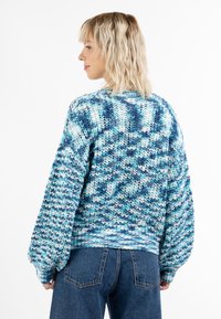 Kötött pulóver, amely kék és türkiz árnyalatok keverékével, oversize fazonnal, bordázott mandzsettákkal és az egész pulóveren végigfutó texturált mintával rendelkezik.