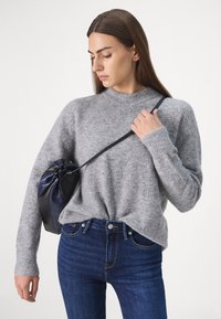 Grå ullgenser med høy krage og ribbestrikkede mansjetter, sammen med mørkeblå jeans. En liten svart snorpose holdes i den ene hånden.