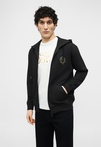 Νεαρός με σγουρά μαλλιά, ντυμένος με μαύρο φούτερ με φερμουάρ πάνω από λευκό T-shirt με χρυσό κείμενο, στέκεται μπροστά σε μια απλή ανοιχτόχρωμη επιφάνεια.