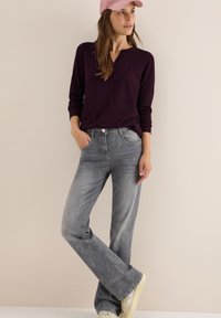 Graue Flared Jeans mit einer verblassten Textur, kombiniert mit einem dunkelvioletten Langarmoberteil und einer pinken Mütze, zeigen ein lässiges Outfit.