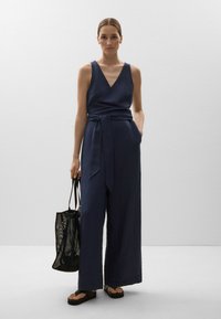 Nachtblaues Leinen-Jumpsuit mit Wickeloptik, weitem Bein und gebundenem Taillengürtel. Kombiniert mit einer schwarzen Netz-Tote-Bag und Flip-Flops.