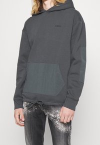 Hoodie grigio scuro con tasca frontale, dettagli testurizzati a contrasto, orlo con cordino e logo del marchio discreto sul petto.