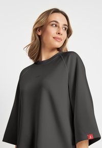 Zwarte oversized t-shirt met driekwart mouwen, raglanontwerp, een zachte textuur en een klein logo op de borst; rode labelaccent op de mouw.