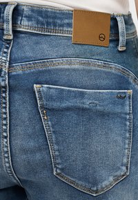 Κοντινή λήψη πίσω τσέπης μπλε τζιν με καφέ δερμάτινο λογότυπο της μάρκας πάνω από την τσέπη και μικρή μεταλλική λεπτομέρεια λογότυπου στη γωνία της τσέπης.