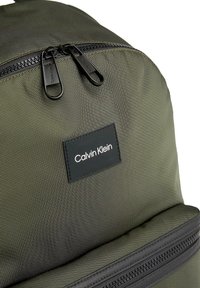 Zaino verde oliva con tessuto testurizzato, chiusura a doppia cerniera e un patch in pelle nera con il logo "Calvin Klein".