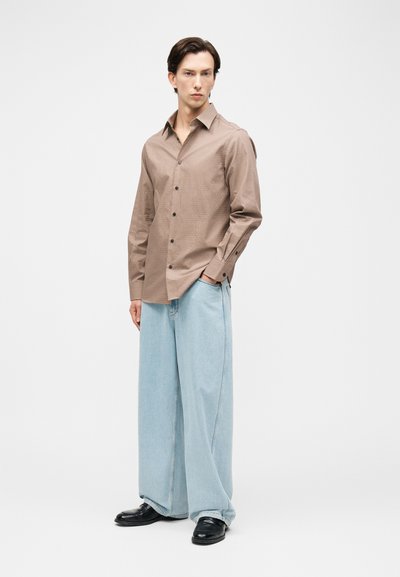 Άνδρας που στέκεται με τα χέρια μερικώς στις τσέπες, φοράει ένα καφέ πουκάμισο με κουμπιά, φαρδιά ανοιχτό μπλε τζιν και μαύρα παπούτσια, σε λευκό φόντο.
