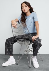Jasnoniebieski T-shirt z białym logo, wzorzyste czarne dżinsy z uśmiechniętymi buźkami i białe trampki, siedzący na metalowym krześle z drutu.