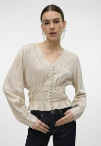 Lichtbeige blouse met een V-hals, geplooide details, knopsluiting aan de voorkant en een gathered taille. Lange mouwen met knoopmanchetten, gecombineerd met donkere jeans.