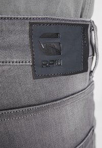 Primer plano de unos jeans de mezclilla gris que muestran un parche de cuero negro cosido con el logotipo en relieve "G RAW" en la cinturilla.