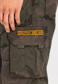 Cargo hlače u tamnozelenoj kamuflažnoj tkanini, s žutim "ALPHA IND." oznakom na prednjem džepu, s vidljivim šavovima i džepovima.