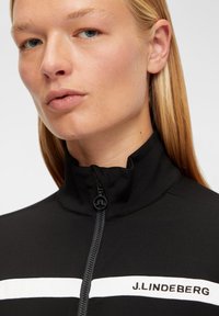 Svart zip-up jacka med hög krage som har vita horisontella ränder och varumärket "J. Lindeberg" i svart på vänster sida.