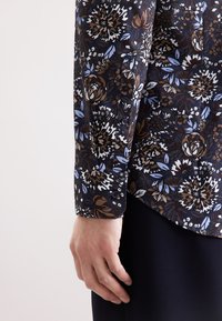 Bloemenpatroon shirt in donkerblauw, met bruine, witte en lichtblauwe bloemen. Heeft knoopsluiting bij de mouwen en een soepele textuur.