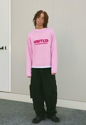 Maglione oversize rosa con testo "UNTLD" e "INTITLED GOODS". Pantaloni cargo neri larghi con tasche. Sneakers nere. Sfondo chiaro.