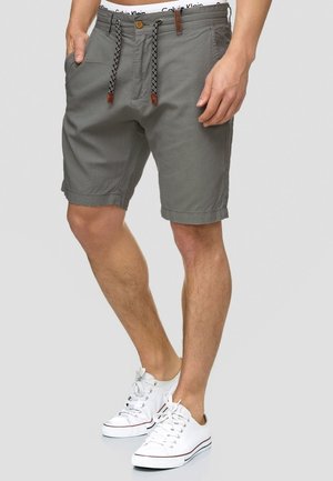 Homme portant un short décontracté gris avec cordon et des baskets blanches, debout avec une main dans la poche sur fond uni.