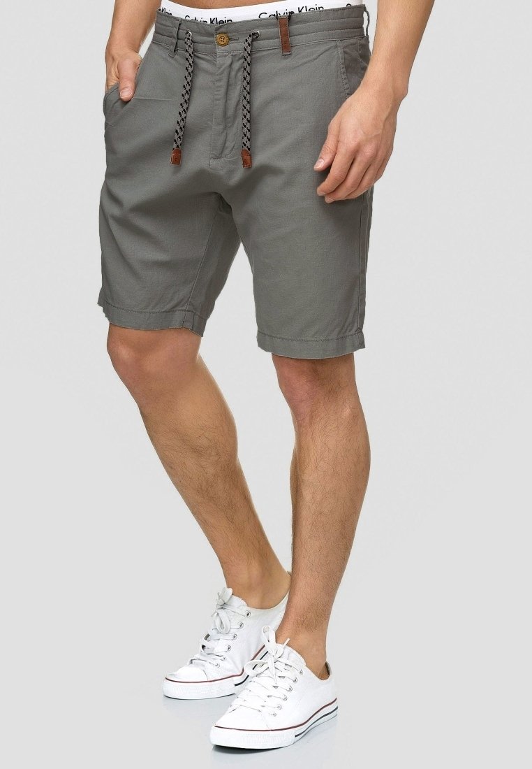 Homme portant un short décontracté gris avec cordon et des baskets blanches, debout avec une main dans la poche sur fond uni.