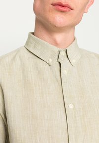 Vaaleanvihreä nappipaita, jossa on jämäkkä kaulus, valmistettu kuvioidusta kankaasta; edessä pieniä helmen kaltaisia nappeja.