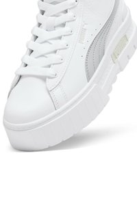 Sneaker in pelle bianca con lacci piatti, suola spessa bianca e una striscia argentata di accento sul lato. Presenta un dettaglio del logo ben visibile.
