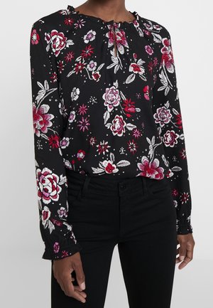 Kvinde iført en sort bluse med lange ærmer med røde, lyserøde og hvide blomsterprint og sorte bukser.