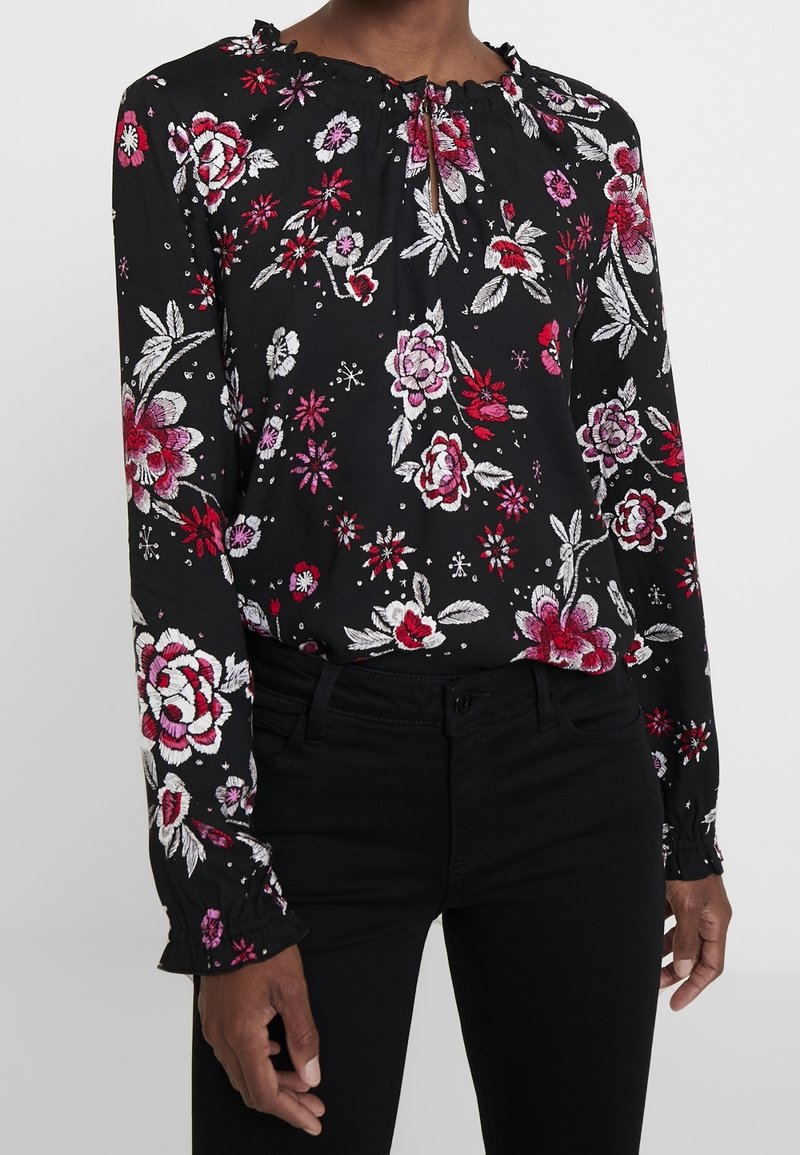 Femme portant un chemisier noir à manches longues avec des motifs floraux rouges, roses et blancs, ainsi qu'un pantalon noir.