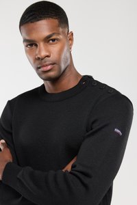 Hombre con cabello corto y pequeño pendiente, vistiendo un suéter negro con botones en el hombro, con los brazos cruzados, fondo neutro.