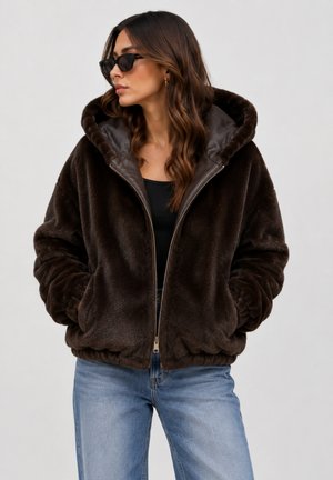 Woman wearing dark brown faux fur hooded jacket, black top, sunglasses, and light blue jeans with hands in pockets facing left.