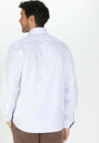 Chemise blanche à manches longues avec un col classique, texture lisse et poignets boutonnés. Présente un ourlet arrondi et aucun motif visible.