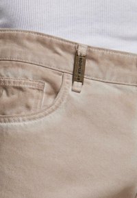 Beige housut, joissa on näkyvät etutaskujen ompeleet ja pieni messinkitagi, jossa lukee "2Y STUDIOS" vyölenkissä, yhdistetty valkoiseen ribbipaitaan.