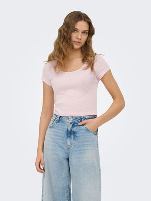Ung kvinde med bølget hår iført en lyserød mønstret kortærmet top og lyseblå højtaljede viddeben jeans.