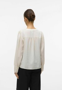 Blouse légère crème avec de longues manches bouffantes, un col arrondi et une texture subtile. Dotée d'une emmanchure dans le dos et de poignets boutonnés. Assortie à un pantalon noir.