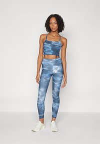 Žena v modrém a stříbrném tie-dye sportovním oblečku, který se skládá z crop topu a legín s vysokým pasem, ve bílých teniskách, stojí na jednobarevném pozadí.