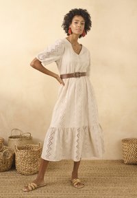 Abito beige al ginocchio con scollo a V, maniche corte a sbuffo e motivi a zigzag traforati. Abbinato con una cintura marrone e sandali piatti.