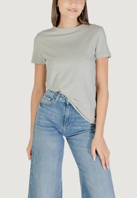 T-shirt in cotone grigio chiaro con maniche corte, caratterizzato da un sottile logo "GUESS". Abbinato a jeans azzurri a vita alta.