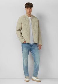 Beige gesteppte Bomberjacke mit Stehkragen, kombiniert mit hellblauen Jeans und weißen Sneakern mit beigen Akzenten. Einfaches und strukturiertes Design.