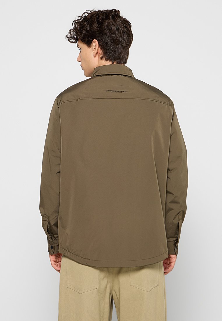 Veste légère marron avec un col, des poignets boutonnés et une coupe droite. Présente un logo au-dessus du dos et du texte sur l'encolure.