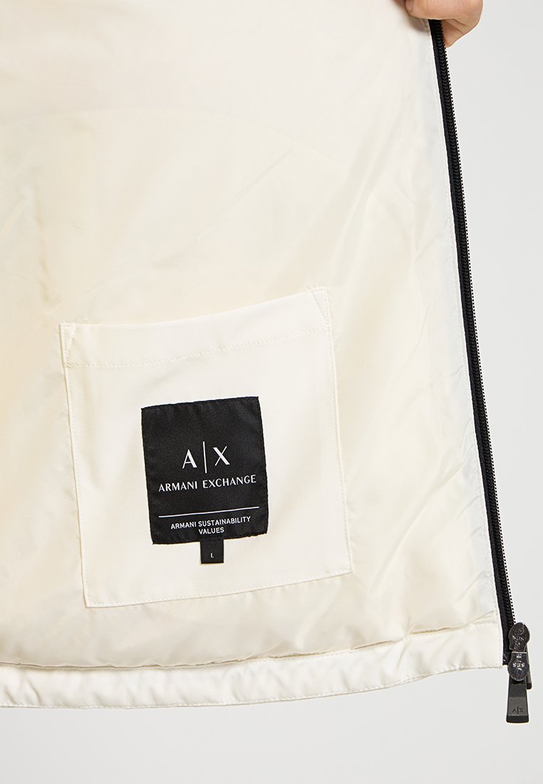 Interno di una giacca di colore crema con un'etichetta nera che riporta "A/X Armani Exchange." Include una piccola tasca e una chiusura con zip.