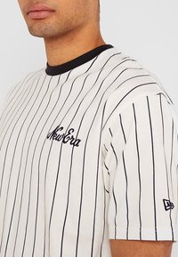 Hombre que lleva una camisa blanca con finas rayas verticales negras, con un collar de canalé negro y el logo de "New Era" en el pecho y la manga.