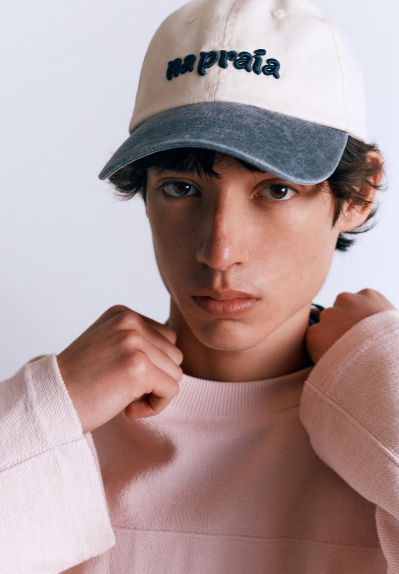 Jeune personne portant une casquette bicolore avec le texte « na praia », ajustant le col d’une chemise rose clair à manches longues, regardant droit devant.