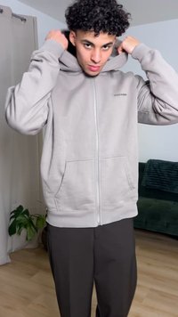 Hellgrauer Kapuzenpullover mit Reißverschluss, Kapuze, Fronttaschen und gerippten Bündchen. Kombiniert mit dunkler Hose, aufgenommen in einem Zimmer mit neutralen Farben.