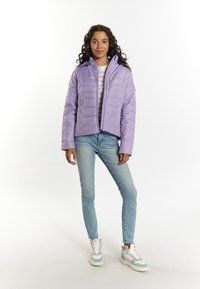 Lavendelfärgad pufferjacka med vadderad textur, dragkedjestängning och sidofickor, kombinerad med ljusblå jeans och flerfärgade sneakers.
