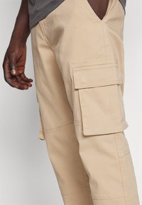 Pantalones cargo beige con un bolsillo lateral, fabricados en tela de algodón con una textura suave. El corte es relajado y casual.