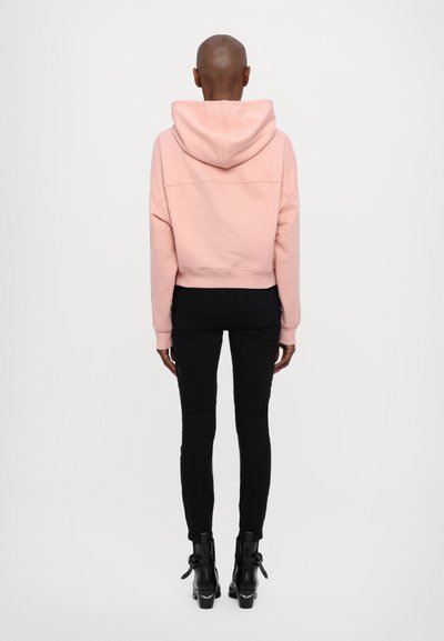 Sweatshirt rosa cropped com capuz, apresentando um corte relaxado e ombros caídos, combinado com jeans skinny pretos e botas de tornozelo.