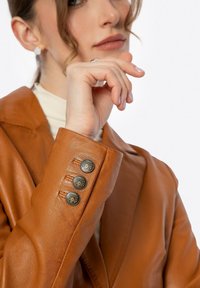 Giacca di pelle marrone con ampi revers, superficie texturizzata e tre bottoni decorativi sui polsini. Vista ravvicinata della mano e dell'accessorio.