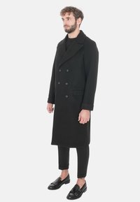 Cappotto in lana nero a doppio petto con ampio colletto a rever, tasche laterali e chiusura a bottoni, indossato sopra pantaloni neri slim-fit e scarpe.