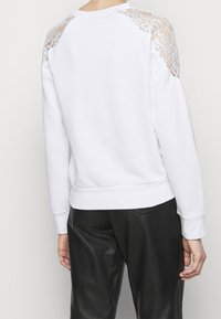 Vit sweatshirt med spetsdetaljer på axlarna, ribbade muddar och nederkant. Slät yta och avslappnad passform, kombineras med svarta läderbyxor.