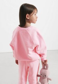 Felpa rosa e pantaloni coordinati in morbido tessuto, con una vestibilità comoda e maniche raglan. Il bambino tiene in mano un giocattolo a forma di coniglio in corduroy beige.