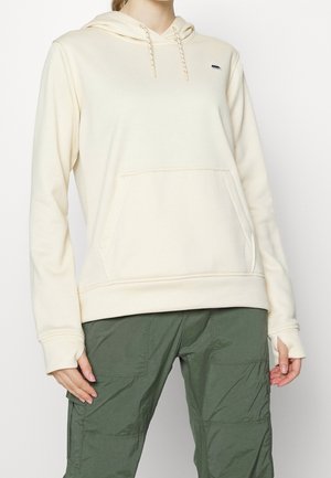 Person iført en cremefarvet hættetrøje med frontlomme og snore, kombineret med olivengrønne cargobukser.